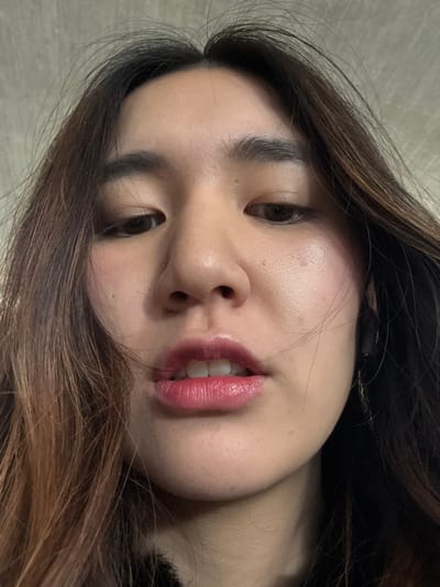 A close-up, slightly rotated shot captures an Asian woman's face. She has fair skin, dark brown eyes, and full, glossy lips painted a pinkish-red hue. Her dark brown hair with lighter highlights frames her face. The background is a neutral, light tone, likely a wall or surface. The lighting is soft and flattering, accentuating her features. The focus is primarily on her face, with the rest of her body and surroundings out of view.