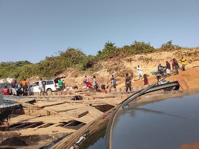 The image depicts a bustling riverine transit point in Ibi, Nigeria, under a clear blue sky. The viewpoint is from the roof of a grey vehicle, with its windshield wiper mechanism visible in the lower right foreground.
In the midground, a substantial wooden platform or ferry deck is constructed from numerous irregularly laid wooden planks. Adjacent to this structure, dark-colored boat hulls are partially visible, one appearing loaded with goods. A red sedan and a white minivan are positioned on or near the wooden platform. Approximately ten individuals are gathered around the white minivan, with its rear hatch open, suggesting active loading or unloading. A separate group of individuals stands near the red vehicle.
To the right and background, a sloping earthen bank of sandy-brown soil rises, topped with dense green vegetation and trees. Approximately 15 to 20 individuals are scattered across this bank. Their attire consists of casual clothing in various colors. Several people are walking, while others stand or interact in small groups. One person is seated on a motorcycle on the upper part of the bank. Another individual, dressed in white traditional attire, stands further back on the slope. The scene conveys a communal hub for transport and commerce.