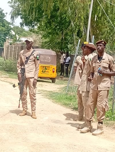 The media file shows a group of uniformed individuals, likely security personnel, standing outdoors in Dutse, Nigeria. The primary subject in the foreground is a man in a light brown uniform, wearing a cap with "NIS" visible and a rifle slung across his body. He appears alert and is looking towards the camera. Behind him, three other men in similar uniforms are standing in a line. The second man from the left is wearing a brown beret, and the third man is also wearing a brown beret. The fourth man, on the far right, is holding a light blue object in his hand.
The scene is a dusty, unpaved area, possibly a roadside or a perimeter. In the background, a yellow auto-rickshaw is parked. Trees and foliage are visible, suggesting an outdoor setting during daylight hours. The sky appears bright, indicating good weather. The uniforms appear to have patches or insignia, but they are not clearly legible in all cases. The overall impression is one of an operational setting where security personnel are on duty. The individuals' postures and the presence of a firearm suggest a situation requiring vigilance, but there are no overt signs of distress or immediate danger. The presence of the auto-rickshaw and the general landscape hint at a location within a populated area in Nigeria.