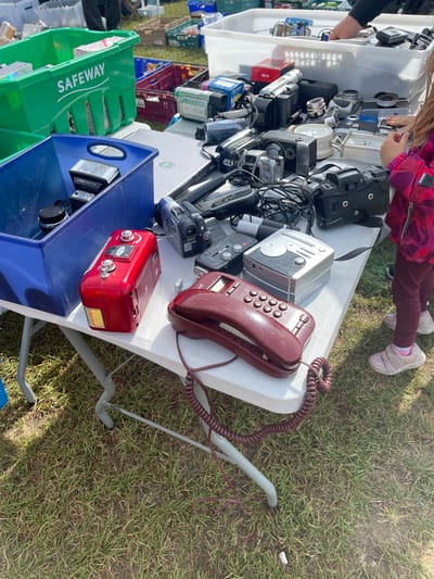 The image depicts a table covered with assorted electronic items, seemingly at an outdoor market or sale. A child is seen examining items on the table. The table displays a variety of devices, including cameras, radios, and a landline telephone. Below the table, crates are used for storage. The location appears to be a grassy area, possibly in the vicinity of Sevenoaks, United Kingdom. A green crate labeled "SAFEWAY" is visible. The objects suggest a collection of vintage or used electronics.