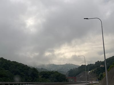The image captures a misty, mountainous landscape near the city of Tsakva, Georgia, under an overcast sky. The foreground features a highway with a concrete barrier and several streetlights. A dark-colored car is visible on the highway, moving away from the camera. The mid-ground consists of lush, dark green trees covering rolling hills. In the distance, the mountains are shrouded in fog, with scattered houses and communication towers dotting the slopes. A red structure, possibly a bridge or crane, is visible on the right side of the highway in the mid-ground. The overall atmosphere is subdued and hazy due to the weather conditions.