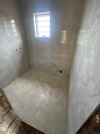 The image shows the interior of a small, unfinished bathroom. The walls, floor, and ceiling are covered in large, light-colored marble tiles. There is a window with bars on the left wall, and the sunlight coming through is reflecting off the tiled floor. There are a few holes in the walls, possibly for plumbing or fixtures. The floor near the edge of the room on the right is unfinished, showing what looks like concrete or brick. The location is Angwan Sayawa, Nigeria.