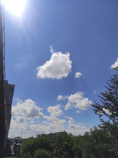 This image depicts a bright, sunny day with a clear blue sky dotted with fluffy white clouds. In the foreground, a portion of a dark, industrial-looking structure, possibly a building or a fence, occupies the left side of the frame. The sun's rays are prominently visible in the upper left corner, creating a lens flare effect. In the middle ground, a dense canopy of lush green trees stretches across the horizon, with glimpses of buildings and a tall tower or pylon peeking through the foliage. A dark-leaved tree branch extends into the upper right corner of the image. The scene appears to be an outdoor urban or industrial environment in Kyzyl, Kazakhstan. There are no people or animals visible in the frame.