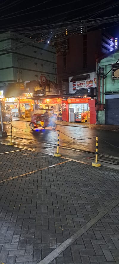 This image depicts a street scene at night in Mandaluyong City, Philippines. The foreground features a textured brick pathway. Beyond this, the street is wet and reflects the neon lights of surrounding establishments. A blurred image of a tricycle, a common mode of transport in the Philippines, is visible moving across the street. Yellow and black striped bollards with chains are placed along the edge of the pathway, indicating a boundary.

In the midground, several food stalls are open for business. One stall has signage that reads "YONG YONG FRIED NOODLE". Another stall features a red facade with "ALL-IN 1 BAKESHOP" written on it, displaying baked goods and pastries inside. Above the stalls, banners advertise "CELEBRATE YOUR PARTY AT KENNY'S". Figures of people are visible, some appearing to be customers or staff at the food establishments.

The background shows the architecture of buildings, some with illuminated windows. Overhead, a dense network of electrical wires crisscrosses the sky. A large banner advertising "22% DISCOUNT" is visible on one of the buildings. The overall atmosphere is that of a bustling urban night scene, characteristic of many commercial areas in Philippine cities.