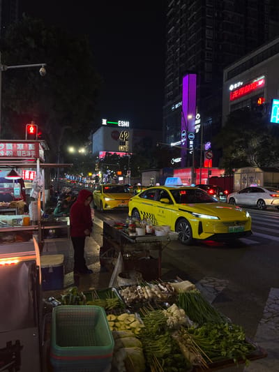 The image captures a vibrant nighttime street scene in an urban area of China, likely in Tian’gongdian Subdistrict. Tall buildings with numerous illuminated signs line the left side of the street, casting a glow on the surroundings.
On the left, several signs are visible, including a prominent purple vertical sign with "KTV" (Karaoke Television), another sign displaying "ESMI," and further down, "无界台政" (Wujie Taizheng), "HOOJA," and "合家" (Hejia) can be seen on various establishments. A red traffic light is visible on a pole near the bottom left.
The street itself is busy with traffic. Several bright yellow electric taxis, a common sight in Chinese cities, are visible. One taxi in the foreground has "A BW9052" on its license plate and the characters "公美" (Gongmei) on its side. Another yellow taxi and a white car are also present on the road.
On the right side of the street, a bustling food stall is set up. It displays a variety of fresh produce, including leafy greens, cabbage, spring onions, and skewers of mushrooms and other vegetables, suggesting a market for street food or ingredients. A person wearing a red hooded jacket stands near the stall, looking towards the camera. Stacked plastic baskets, likely for produce, are also visible next to the stall. The atmosphere is typical of an active urban night, with commercial activity and people out on the streets.
Visible text from the scene includes "沙面凉面" (Shamian Cold Noodles) and "少粉锅巴洋芋" (Less flour crispy rice potato), which appear to be food items offered at another stall or nearby establishment.