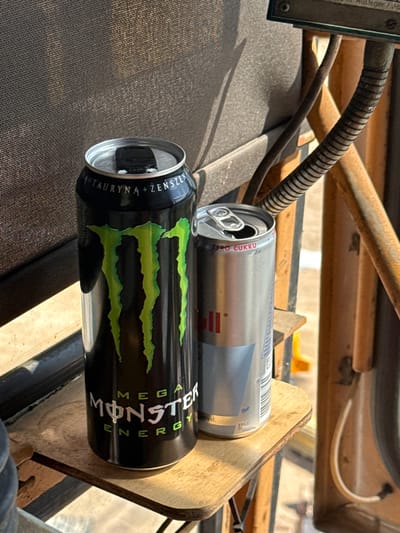 The image shows two cans of energy drinks placed on a textured, dark surface, possibly a seat or fabric covering. The can in the foreground is a black Monster Energy can with the distinctive green claw mark logo and white text reading "MEGA MONSTER ENERGY". The text on the side of the can includes "TAURYNA + ŻEŃ-SZEŃ" in white lettering. The can in the background is a silver can, possibly a Red Bull, with a white tab. Text on this can includes "ZERO CUKRU". Both cans appear to be open. The cans are resting on a wooden surface. In the background, there are metal and wooden structures, suggesting an outdoor or industrial setting. A corrugated tube and a thin wire are visible, adding to the sense of a functional or mechanical environment. The lighting suggests it is daytime, with sunlight casting shadows. There are no people visible in the image. The scene appears to be in Mikołajki, Poland.