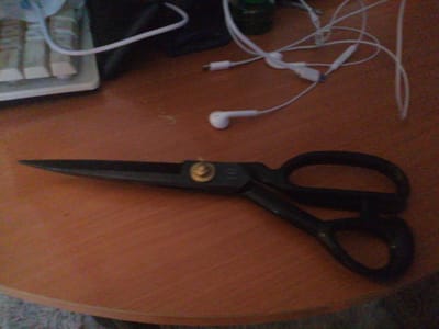 In the city of Степной, Russia, a pair of black-handled scissors with a gold pivot lies on a wooden table. The scissors are positioned horizontally across the frame, with the blades pointing left. To the upper right of the scissors, a tangle of white wires and earphones is visible, suggesting a workspace or desk area. A portion of a keyboard with white keys and a light-colored surface is visible in the upper left corner of the image. The overall lighting is dim, casting shadows and giving the scene a muted tone.