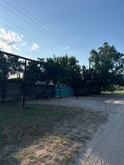 The image depicts a rural scene in Baidivka, Ukraine, under a clear blue sky with scattered clouds. In the foreground, a dirt road with tire tracks curves to the right. To the left of the road is a patch of dry grass and earth, transitioning into a more sparse grassy area with some weeds. Beyond this, a horizontal metal pipe supported by vertical posts stretches across the midground. Behind the pipe, a concrete wall runs horizontally, separating the area from trees and a turquoise gate. Several trees of varying sizes with dense foliage are visible behind the wall and gate, with larger trees dominating the right side of the frame. The overall impression is one of a quiet, possibly residential, outdoor area in a village setting.
