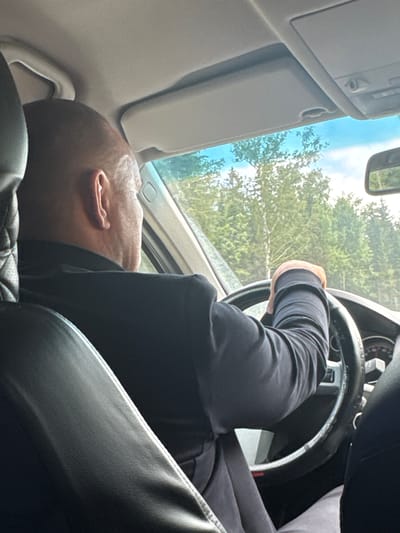 The image shows a man driving a car. He is bald and wearing a black jacket. His hands are on the steering wheel. The car appears to be moving on a road surrounded by trees. The time of day appears to be daytime with sunlight visible. The image is taken from inside the car, looking over the driver's shoulder. The car's interior is visible, including the steering wheel and dashboard. There are no specific details in the image that indicate the location is in Didino, Russia.