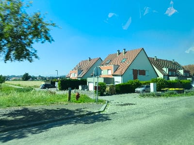 A neighborhood with houses with orange-tiled roofs under a bright blue sky. A field of farmland is visible in the background, along with a few trees. The houses are surrounded by green hedges and some cars parked in front. In the foreground is a road and some grass on the side. A fire hydrant is visible in front of one of the houses.