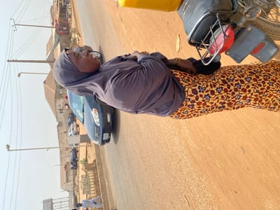 This image depicts an outdoor scene on a street in Lapai, Nigeria, under clear daylight conditions.
A female individual of dark complexion is the central subject, positioned in the mid-ground, facing towards the viewer's left. She wears a grey head covering (hijab), a matching long-sleeved top, and a long skirt or wrapper featuring a yellow-brown background with blue and reddish-brown floral patterns. Gold-colored bangles are visible on her right wrist. Her expression is neutral, and she is looking slightly upwards and to her right.
To the immediate right of the female subject, the rear section of a black motorcycle is visible, equipped with a red taillight and a license plate bracket. A yellow jerrycan is partially seen at the top right, possibly secured to the motorcycle. Behind the woman, a dark blue sedan, consistent with a Toyota Corolla model, is present, either parked or moving slowly along the reddish-brown road. Further down the street, additional vehicles, including cars and a white van, are parked along the roadside.
The environment includes a reddish-brown road, utility poles with multiple wires, and streetlights lining the thoroughfare. Various single-story or low-rise buildings with brown roofs are situated along both sides of the street in the background. Faint details in the far background suggest commercial activity or storage near some structures. A person in a white garment is partially visible on the far left, near the bottom edge of the frame.