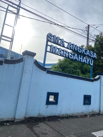 The image shows a blue and white wall with a sign that reads "SMA ANGKASA MARGAHAYU". The wall is located in Margahayu, Indonesia, at the coordinates -6.982, 107.563. The wall is topped with a brown molding and has two square, dark blue openings. There is a sidewalk in front of the wall, and several power lines are visible in the sky. The sky is overcast. The sign is mounted on a blue pole. There are no people visible in the image. The wall appears to be part of the entrance to the "SMA ANGKASA MARGAHAYU", which is likely a school.
