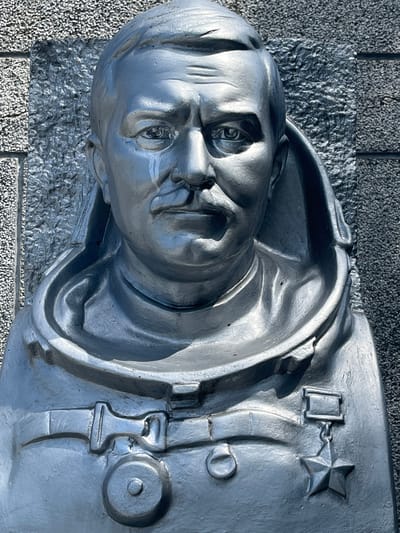 This media file is a close-up, low-angle shot of a silver-colored bas-relief sculpture of a person's head and upper torso. The sculpture is mounted on a textured stone surface, possibly a wall or monument. The subject's face is detailed, with a stern expression, closed eyes, and prominent features. They are depicted wearing a uniform with what appears to be a star-shaped medal or insignia on the chest, attached by a chain to a rectangular piece of metal. There are also other uniform details visible, such as a buckle and what looks like a button. The lighting suggests it is daytime, with the sun casting shadows that highlight the contours of the sculpture. The background consists of a coarse, gray, pebble-like texture. The overall impression is one of solemnity and remembrance, possibly commemorating a historical figure or event. The setting appears to be outdoors in a public space. There is no visible text in the image.