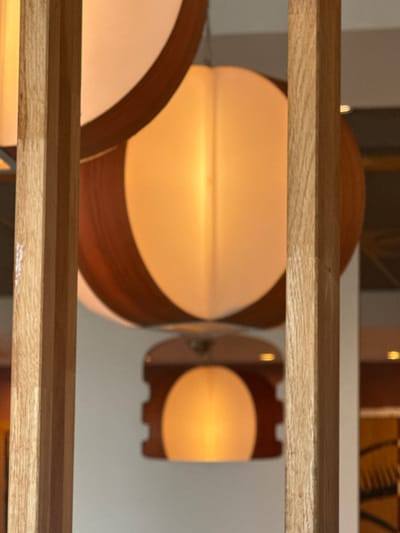 The image shows a close-up of a decorative room divider made of vertical wooden beams. Behind the divider, two lamps are visible. The larger lamp in the foreground has a round, cream-colored shade with dark wood accents. The smaller lamp in the background is also round and appears to be emitting a warm, yellow light, framed by a dark wood fixture with decorative cutouts. The overall lighting suggests an indoor setting, possibly a restaurant or lounge. The image does not contain any people or identifiable landmarks of Oosterhout, Netherlands.