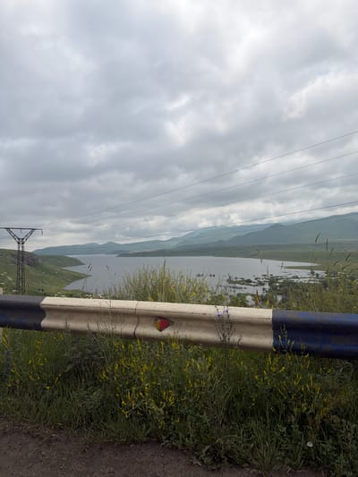 The image depicts a scenic landscape with a body of water, rolling hills, and a cloudy sky. In the foreground, there is a roadside guardrail. To the left, a large power line tower is visible. The environment suggests a rural or mountainous area. The location is identified as Tsghuk, Armenia.