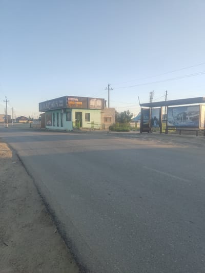 The image depicts a street scene in the city of Aral, Kazakhstan, under a clear, pale blue sky. The focal point is a light green building with a dark sign that reads "MARVANA Tasty Food" in large letters, with "FAST FOOD" and "MILK SHAKE ICE CREAM" also visible. The building appears to be a commercial establishment, possibly a cafe or restaurant, with windows along its front. To the right of the building, there is a bus stop shelter with advertisements displayed. The street in the foreground is paved with asphalt, and there is a dirt shoulder on the left side. Power lines and utility poles are visible in the background, suggesting an urban or semi-urban environment. The overall impression is of a quiet, daytime scene in a relatively modest commercial area.