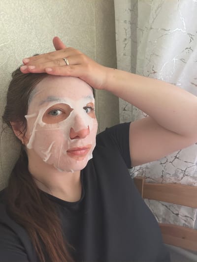 A person is lying on a light-colored surface, possibly a floor, wearing a black garment and a facial mask. The person's head is tilted slightly to the right. Their right hand is raised, resting on the head, with a ring visible on one finger. Brown hair is visible near the bottom of the frame. Part of a light-colored patterned surface is visible behind their arm. A wooden object, possibly furniture, is also visible in the background. The location in the city of Tokkari, Russia, cannot be confirmed from the image.