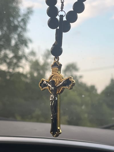 The image shows a close-up of a crucifix pendant hanging from a string of dark beads, likely a rosary. The crucifix itself is detailed, with a depiction of Jesus Christ and the inscription "INRI" at the top. The pendant has a metallic sheen, appearing to be gold and black. The background is blurred, showing greenery and a sky with soft clouds, suggesting an outdoor setting, possibly viewed through a window. The foreground is dark and out of focus, hinting at the interior of a vehicle. The location of Никольское, Russia is not identifiable from the visual information provided in the image.