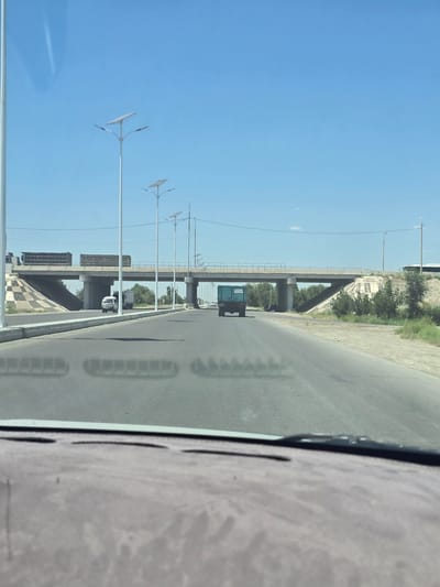 The image captures a view from inside a vehicle looking out at a highway with a bridge overhead. The sky is clear and blue with a few wispy clouds. Several modern streetlights with solar panels are visible along the side of the road. On the highway, a small white van and a dark green truck are driving. The bridge spans the highway, and two large trucks are seen driving on it. The surroundings appear to be a dry, somewhat arid landscape with sparse vegetation. The scene is likely in Dehcha, Uzbekistan, given the overall environment and infrastructure.