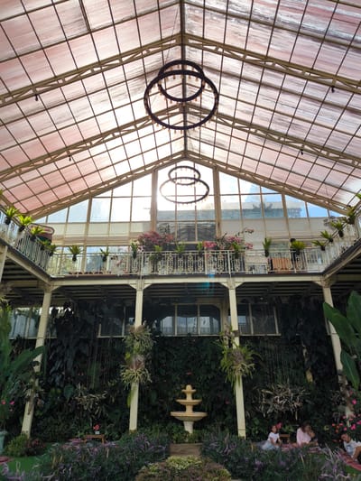 The image captures an interior view of a glasshouse or conservatory in Candikuning, Indonesia. The structure features a high, geometrically-patterned roof supported by metal beams, with two circular chandeliers hanging from the ceiling. A second-story balcony adorned with plants and flowers overlooks the ground floor. The ground floor is densely populated with greenery, including a variety of plants and a tiered fountain. Several people are seated on the ground among the plants, engaging in conversation or relaxation. The overall atmosphere is lush and serene.
