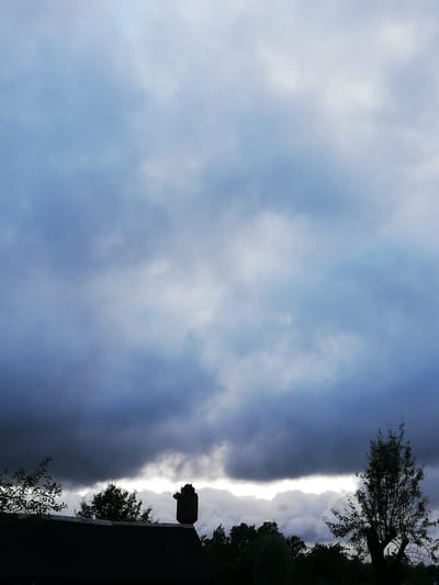 The image depicts a dark, overcast sky with heavy clouds over the landscape of Novapahostski Sel'ski Sovet, Belarus. In the foreground, the silhouetted roof of a building is visible, with a chimney structure on top. To the left and right of the roofline are silhouettes of trees and foliage. A single tree with sparse leaves stands prominently on the right side of the frame. The lighting suggests it is either early morning or late evening, with a bright band of light breaking through the clouds along the horizon.