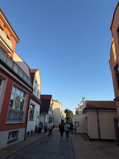 This is a sunny day photograph of a narrow cobblestone street in the historic district of Rostock, Germany, likely near the harbor area given the traditional architecture. On the left, a building with a vibrant orange facade and white trim features a balcony with a decorative metal railing and flower boxes. To its right, a white building with a tiled red roof and a modern window is visible. The street is lined with several other buildings, most with tiled roofs, in varying shades of white, yellow, and red.
Several pedestrians are walking along the street. In the foreground, on the right sidewalk, a man and a woman wearing matching blue and white patterned shirts are walking away from the camera. Further down the street, a group of people are walking in both directions, some carrying bags. The sky is a clear, bright blue, indicating good weather. The lighting suggests it is late afternoon or early evening, casting long shadows and highlighting the warm tones of the buildings. The scene depicts a lively, tourist-friendly atmosphere.