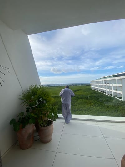 A man in pajamas is standing on a white tiled balcony overlooking a green landscape that may be a mangrove or lagoon. The man's back is to the viewer. In the foreground are two potted plants. In the distance are buildings along the coast. The view is from El Caracol, Mexico.