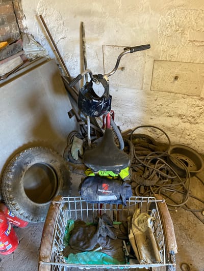 The image shows a cluttered storage area with a light-colored, rough-textured wall. In the foreground, a black bicycle seat is mounted on a bicycle frame. Attached to the handlebars is a black helmet with white accents. Various items are piled around the bicycle, including a black bag with the text "SECURE" and a yellow garment, a stack of flat grey materials, and a worn tire. To the right, a metal wire rack holds several pairs of work gloves and some green plastic sheeting. Behind the bicycle and rack, a large tangle of dark cables or hoses is coiled. A rusty metal rim is also visible. The lighting suggests it could be daytime, but the enclosed space with artificial lighting is not clear. The overall impression is of a disused or storage area for tools and equipment, possibly in a workshop or garage. There is no indication of people or any specific event taking place. The location context provided is "Police, Poland", but there are no visual cues in the image to confirm this.