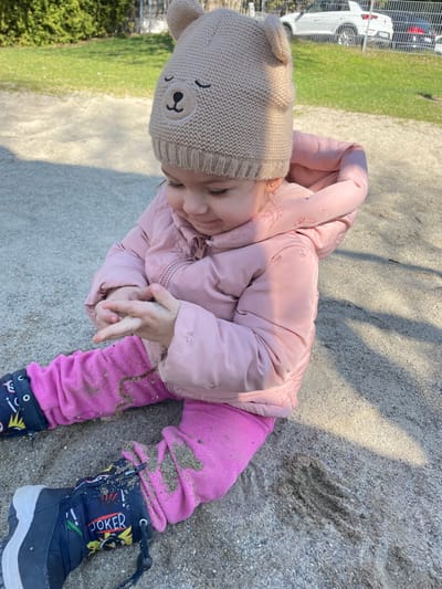 A young child, presumably in Senden, Germany, is seated in a sandpit, observed from a slightly elevated perspective. The child wears a light beige knitted beanie with embroidered sleeping bear facial features and small ears, a light pink padded jacket, fuchsia pants, and dark blue boots decorated with "JOKER" and other graphic motifs. Sand is present on the child's hands, which are clasped together, and scattered across their pants and boots. The child's gaze is directed downwards towards their hands. The immediate environment consists of sand. In the background, green grass and foliage are visible to the left. To the right and further back, a light-colored SUV is parked or situated on a paved surface, with additional vehicles partially visible behind it, bordering what appears to be a public area or park. Distinct shadows indicate direct sunlight.