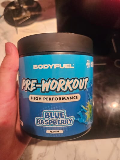 The image shows a close-up of a blue plastic container of "BODYFUEL PRE-WORKOUT HIGH PERFORMANCE BLUE RASPBERRY FLAVOUR!" The container has a black lid and is held by a hand with visible fingernails. The background is blurred and appears to be a countertop with some out-of-focus objects. There is no information in the image or the provided crops that indicates this scene is located in Versailles, France, or any specific city. The image focuses solely on the product.