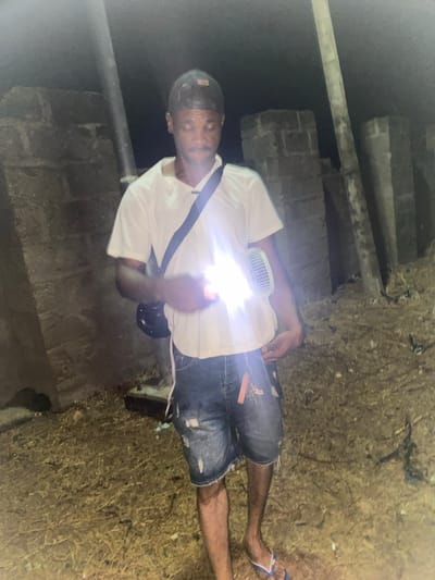 A man stands on what appears to be a dirt road or ground in Katsina Ala, Nigeria. He is wearing a white T-shirt, dark denim shorts, and flip-flops. A strap crosses his chest. He is holding a light. The environment seems dimly lit. There is a structure made of stones in the background, on the left. To the right is ground covered in a thatch-like substance.