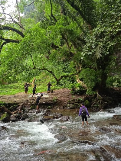 The image shows a group of people standing and walking in a shallow river in Tawangmangu, Indonesia. The river is rocky and has rapids flowing over the rocks. The people are standing on the rocks, and some are walking across the river. There is dense green vegetation and trees on both sides of the river. The coordinates -7.654, 111.136 are approximately in the area of Tawangmangu, Indonesia. One person is wearing a purple jacket, a black backpack, and black shorts. Another person is wearing a blue shirt and black shorts. Another person is wearing a red shirt and black shorts. Another person is wearing a black shirt and black shorts. Another person is wearing a black shirt and black shorts. Another person is wearing a white shirt and black shorts. Another person is wearing a white shirt and black shorts. There is a white blanket and some belongings lying on the ground next to the river. The image was taken from a vantage point overlooking the river.