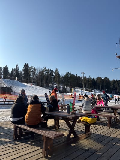 The image depicts an outdoor winter scene at a ski resort located in Vasil'evo, Russia, under a clear blue sky. In the foreground, a wooden deck supports multiple dark brown wooden picnic tables with attached benches. Four individuals are seated at the leftmost table: one person in a dark jacket, one person in a light grey jacket and hat, one person in an orange quilted vest, and another person in a dark jacket. Items on this table include a dark thermos, a green bottle, and a metal cup. Two additional individuals are seated at an adjacent table to the right: one female in a grey puffer jacket and another female in a pink ski jacket and pants. A yellow bag is also visible on their table. Skis and snowboards are propped upright near the tables. In the mid-ground, several individuals are standing or moving on a snow-covered area, some equipped with skis. A person in an orange jacket and red helmet stands near a pair of skis. The background reveals a snow-covered ski slope flanked by dense evergreen forests. Orange and yellow safety netting lines parts of the slope, and ski lift infrastructure is visible. On the far right, a pole structure supports antennas and a CCTV camera. Sunlight illuminates the scene, casting shadows on the wooden deck.