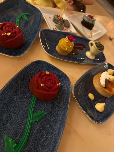 This image captures a dining table laden with multiple plated desserts, consistent with the Be Our Guest restaurant located within Magic Kingdom in Bay Lake, Florida, United States. In the foreground and mid-ground, two dark blue, oblong, textured plates each display a red, rose-shaped dessert with a flocked surface and gold leaf accents, presented on a molded green stem and leaf design. Adjacent, a third dark blue, oblong plate features a yellow, ridged dessert resembling a teacup, topped with a small red flower and white edible pearls, positioned next to a white, tilted, cup-shaped dessert with green frosting and gold flakes. This plate also includes a red sauce drizzle and small white edible spheres. On white, rectangular plates in the background are three gray, spherical cake pops or truffles adorned with multicolored sprinkles, and a dark square cake topped with light green frosting and a red flower. A separate dark blue, square-shaped plate holds a round pastry topped with white whipped cream, nuts, and a dark seed, accompanied by two dollops of yellow cream or sauce. The table surface is uniform light brown. In the out-of-focus background, partial views of two individuals, one wearing a yellow garment, are discernible.