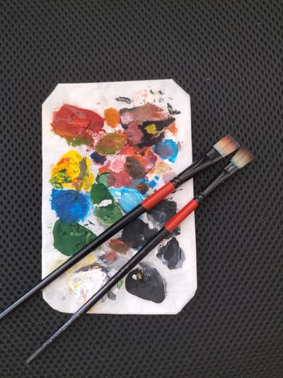 The image shows a palette with an assortment of colorful paints, including red, yellow, blue, green, and various shades of brown and pink. Two paintbrushes with black handles and red accents are placed on top of the palette, with their bristles resting on the paint. The palette itself appears to be made of paper or a similar absorbent material, with dried paint splatters around the edges. The background is a dark, textured surface, possibly a fabric or a mat. The lighting suggests an indoor setting, likely a studio or art space. No specific time of day or weather conditions can be inferred. The scene is static, showing art supplies ready for use or recently used. There is no visible text in the image.