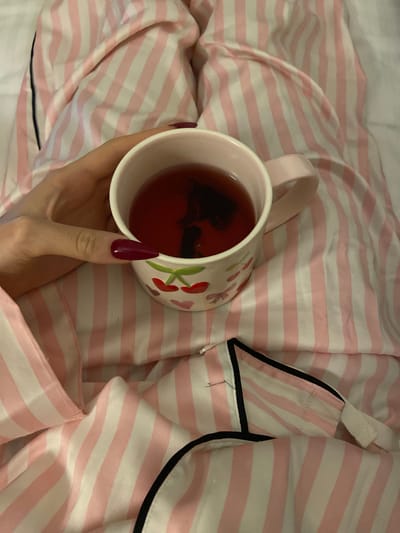 A hand holds a mug of dark liquid, possibly tea, against a background of pink and white striped fabric, likely pajamas. The mug has a floral and cherry design. The hand has burgundy-colored, elongated nails. The pajamas have black piping along the edges and a white shoulder strap. The scene is located in Business Bay, United Arab Emirates.