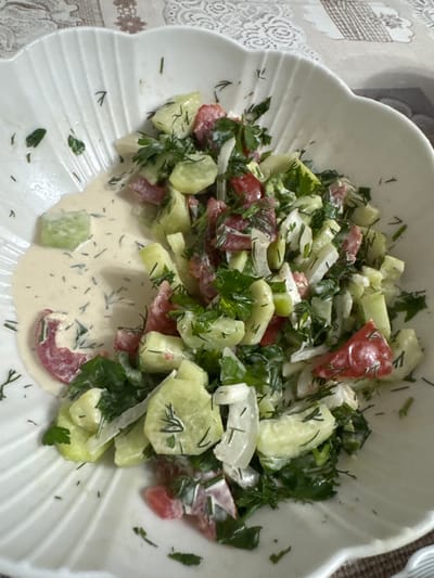 A close-up shot captures a white, fluted bowl filled with a fresh salad. The salad is composed of chopped cucumbers, tomatoes, and onions, all tossed in a creamy white dressing and garnished with fresh dill and parsley. A single slice of cucumber sits apart from the main salad in a pool of the dressing on the left side of the bowl. The bowl rests on a patterned tablecloth. The image appears to be taken indoors, likely in a dining setting in Таганрогский, Russia.