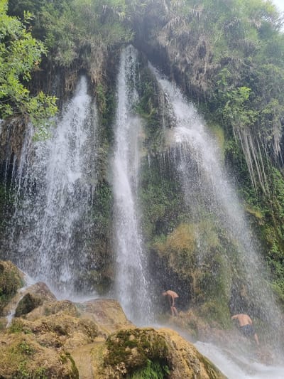 A wide shot captures a cascading waterfall at the base of a lush, green cliff face. Two young men, one in the foreground and one slightly behind him, are positioned near the base of the waterfall, partially obscured by the spray. The man in the foreground, facing away from the camera and to the right, appears to be wearing dark shorts. The second man, to the left, is also facing away from the camera and is bent over. The waterfall itself is dense and powerful, with water falling in many streams and sheets, creating a misty environment. The cliff is covered in green vegetation and moss, with some rocky outcrops at the bottom. The overall scene suggests a natural, outdoor location, possibly a popular spot for swimming or recreation. The location of this waterfall is in Anna, Spain.