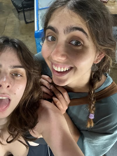 Two young females are depicted in a close-up, informal photograph. The prominent female, positioned right of center, has dark brown hair, which includes a braid secured with a purple elastic on the visible side. She has prominent eyebrows, green eyes, and a broad smile revealing upper teeth. She wears a light teal shirt with a brown accent and two small, intricate silver-toned earrings. Her hands are clasped near the shoulder of the second female. The second female, partially visible on the left, has dark brown wavy hair, her mouth open with her tongue slightly extended, and visible dental braces on her upper teeth. She wears a dark-colored top. The background is out of focus, showing indistinct elements including a light brown surface, a dark object, and a segment of a blue and white structure, consistent with an indoor or covered outdoor setting. Lighting appears artificial. The image contains no identifiable landmarks or specific features to determine a precise location within Thap Sakae, Thailand.