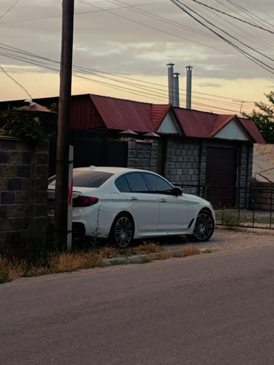 A white sedan is parked on the side of a paved road. The car is positioned next to a brick wall and dry grass. Behind the car is a building with a red roof and stone walls. Three tall cylindrical chimneys rise from the roof of the building. Power lines are visible in the sky, which is overcast with soft yellow and grey tones, suggesting either sunrise or sunset. A wooden utility pole stands to the left of the car, partially obscuring it. The scene appears to be in Orto-Sai, Kyrgyzstan, with a slightly rural or suburban feel. There is no discernible activity taking place, as the car is parked and the scene is still. The lighting and cloudy sky create a muted, tranquil atmosphere. No visible text is present.