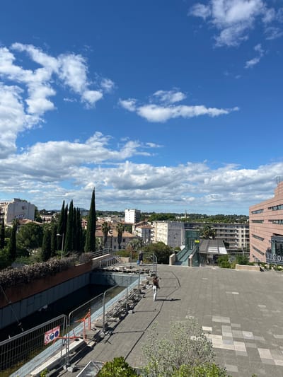 The image is an elevated view of Montpellier, France. The sky is predominantly blue with scattered white clouds. The foreground shows a paved area with a metal fence and scaffolding along the edge. Beyond the fence, a dense row of tall, thin cypress trees stretches across the midground. Buildings of varying heights and architectural styles are visible throughout the cityscape, with some modern, pink-toned buildings located at the upper edge of the frame. A person is walking on the paved area. A sign is attached to the scaffolding.