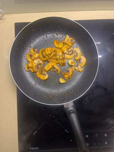 In a kitchen located in Pedrouços, Portugal, a black non-stick frying pan rests on a black TEKA electric cooktop. The pan contains approximately twenty to twenty-five sliced button mushrooms and five to seven whole or halved garlic cloves. The mushrooms exhibit a cooked, slightly browned appearance and are uniformly coated in a yellowish-orange sauce with visible dark green and reddish seasoning specks. Residual oil and sauce are visible on the interior surface of the pan. The electric cooktop features faint white markings for cooking zones and control icons. The cooktop is integrated into a light beige countertop, visible on the left and upper edges of the frame. No individuals are present in the image.