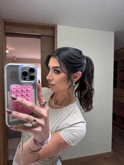 An image depicts a woman taking a mirror selfie in an indoor residential setting. The woman, with dark hair styled in a high ponytail and loose facial framing strands, wears a light-colored, short-sleeved top with ruched detailing. Her right hand, with dark-painted nails, holds an iPhone 14 Pro/Max in a clear case, featuring a religious figure sticker (Virgin Mary/saint) and a pink pop-it grip. This hand displays multiple rings, including a prominent green emerald-cut or oval-cut stone ring on the middle finger, a gold-toned band, and another gold-toned ring on the ring finger. Her right wrist is adorned with two gold-toned bangles and a purple beaded bracelet. She wears a silver-toned necklace with small, clear stones and a matching emerald-cut or oval-cut green stone drop earring in her left ear, accompanied by a small stud. Her makeup includes green eyeshadow and defined eyebrows.
The background, largely a reflection in the mirror, shows an interior space with light-colored walls. To the left, a wooden framed doorway leads to another room with a visible ceiling light fixture. To the right of the woman, a light beige wall transitions to a partial view of a wooden built-in shelving or wardrobe unit, suggesting a dressing area or walk-in closet. The floor visible at the bottom of the image is light wood. The specific city location of San Luis Potosí City, Mexico, is not discernible from the visual information provided in the image.