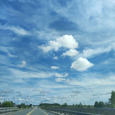 The image depicts a highway scene in Putkovskaya, Russia, under a partly cloudy sky. A two-lane asphalt road with yellow dividing lines stretches into the distance, bordered by metal guardrails. On the left side of the road, there is a green wooded area with some buildings and power lines visible. On the right side, more trees and distant structures can be seen. A white truck and a car are traveling on the highway. The sky is filled with a mixture of white cumulus clouds and thinner, wispy cirrus clouds against a vibrant blue backdrop.