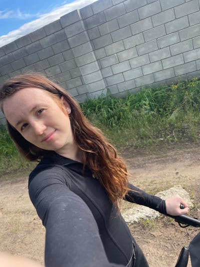 This image, captured in Vasilevka, Russia, features a female subject taking a selfie outdoors. The subject, appearing to be in her 20s or early 30s, has long, wavy reddish-brown hair and a light complexion with visible freckles. She is wearing a black long-sleeved top with a partial zipper closure at the neck. A small, silver-toned, snowflake-shaped stud earring is present in her right earlobe. She looks directly at the camera with a slight, closed-mouth smile. Her right hand is extended towards the camera, gripping a black handle, which is part of an unseen object, possibly a bicycle or stroller, partially visible in the lower right corner. The background consists of a grey concrete block wall running diagonally from the upper left. At the base of the wall and along the foreground, there is green grass mixed with small yellow wildflowers. The ground in the foreground and midground is an unpaved dirt path. A segment of blue sky with white clouds is visible in the upper left.