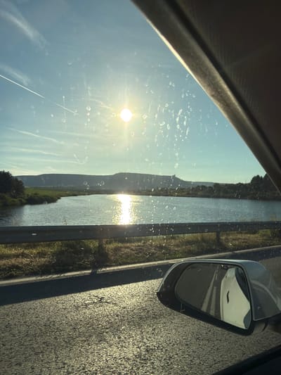 The image shows a view from inside a car, looking out the passenger side window. The sun is shining brightly in a clear blue sky, with some faint clouds and a contrail from an airplane visible. The car window has water spots or dirt on it, distorting the view slightly.

In the distance, there is a body of water, likely a river or lake, with its surface reflecting the sunlight. On the far side of the water, there are rolling hills covered in vegetation. The immediate foreground shows a road with a guardrail separating it from the water and embankment. A side-view mirror of the car is visible in the lower right portion of the image, reflecting the road and sky. The scene appears to be set in a rural or semi-rural environment, possibly near the city of Dibich, Bulgaria, given the landscape and the presence of a road and body of water. There are no people or specific objects of interest clearly identifiable in the scene other than the general landscape and elements of the car.