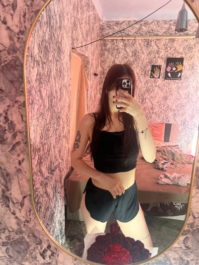 In the city of Blizhnii Khutor, Moldova, a young woman with long brown hair is captured in a mirror selfie. She is wearing a black crop top and black shorts. Her right arm, visible in the reflection, has a tattoo of a dragon on her upper arm and a delicate bracelet with a charm on her wrist. Her left hand is holding a smartphone, which she is using to take the photo.

The background of the reflection shows a bedroom. The walls are covered in a textured, marble-like wallpaper with pink and gray tones. A bed with rumpled blankets is visible, along with some clothing items scattered on top. There are two framed handprint artworks hanging on the wall behind the bed. In the foreground of the reflection, a bouquet of deep red roses is placed in front of the mirror. The room is dimly lit, with a pinkish hue cast by an unseen light source, possibly from the left side of the image. The mirror itself has a wide, oval, gold-colored frame.