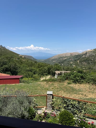 This image depicts a scenic landscape with a view of the sea and hills, located in Mikros Prinos, Greece. In the foreground, there is a wooden railing with stone pillars, a garden with lavender, flowers, and greenery, and the roof of a red building is visible on the left. Beyond the garden, the terrain slopes upwards, covered with trees and vegetation. A structure with wooden beams and concrete supports is under construction in the midground, partially obscured by foliage. In the distance, rolling hills dominate the landscape, with a body of water, likely the sea, and distant islands or a mainland coastline under a bright blue sky with scattered white clouds. The overall scene suggests a rural or semi-rural setting in Greece during a sunny day.