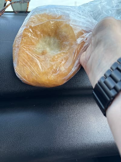 A close-up, high-angle shot shows a fried dough pastry, likely a pirog or similar baked good, sealed in a clear plastic bag. The pastry is golden brown and appears slightly greasy, with a textured surface. A person's hand, wearing a dark, segmented watch, is visible on the right side of the frame, holding the bag. In the upper left corner, a pair of glasses with dark tortoiseshell frames rests on a dark, textured surface, possibly a car dashboard. The background is a dark, textured surface, consistent with the interior of a vehicle. There are no discernible urban elements or landmarks that would indicate a specific location within Chernogorsk, Russia.