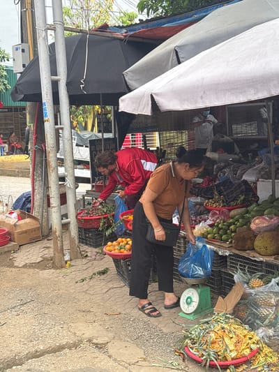 In Bắc Yên, Vietnam, an outdoor market scene unfolds under a covered stall. A man wearing a red jacket and dark pants is selecting fruit from a red basket. To his right, a woman in a brown shirt and black pants is weighing fruit in a blue plastic bag on a green scale. The market stall displays a variety of produce including oranges, plums, avocados, and jackfruit. In the background, a few people are visible, and the market appears to be situated along a street.